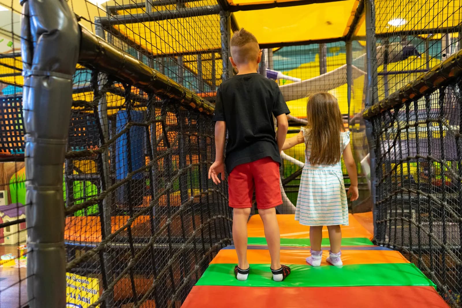 Due bambini camminano di spalle sul ponte colorato del playground alto sette metri